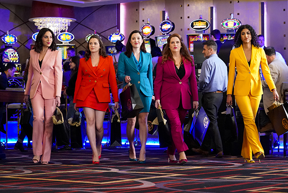 Five women in bright pink, red, teal, magenta, and yellow suits walk through a casino carrying shopping bags.