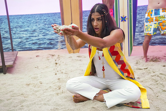 A woman in white pants and a colorful vest sits in sand, letting sand pour from her hands.
