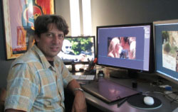A man in a plaid shirt sits at a desk with two computer monitors displaying images.