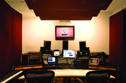 A recording studio control room featuring multiple computer monitors, studio speakers, and audio mixing equipment.