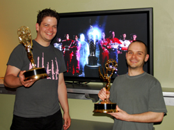 Two smiling men standing in front of a TV, each holding a gold trophy.