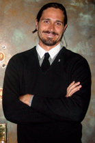 A smiling man with dark hair and a goatee wearing a black sweater, white shirt, and black tie.