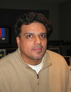 A man with dark curly hair and a goatee wearing a tan quarter-zip fleece jacket.
