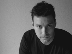 Black and white portrait of a man in a dark t-shirt looking directly at the camera.