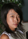 A woman with dark, shoulder-length hair looks toward the camera against a background of greenery.