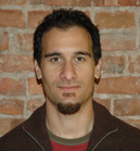 A man with dark hair and a goatee looking at the camera against a red brick wall.