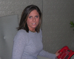 A woman in a grey sweater sits in a chair and holds a red landline telephone.