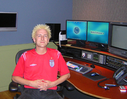 A man in a red shirt sits in a chair before a desk with multiple computer monitors and equipment.