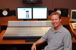 A man smiling while sitting in front of a large mixing console and dual computer monitors in a studio.