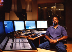 A man sits in a dark recording studio surrounded by computer monitors and audio mixing consoles.