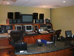 A control room with multiple computer monitors, speakers, and workstations on wooden desks.