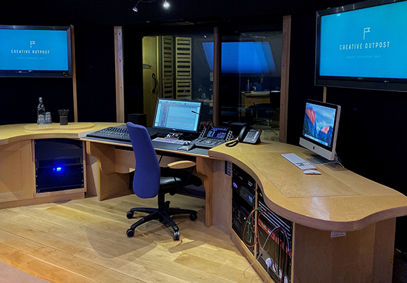 A curved wooden workstation in a dark studio featuring computer monitors, audio equipment, and a blue office chair.