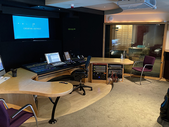 Audio production studio featuring a large mixing console, computer monitors, and curved wooden workstations.