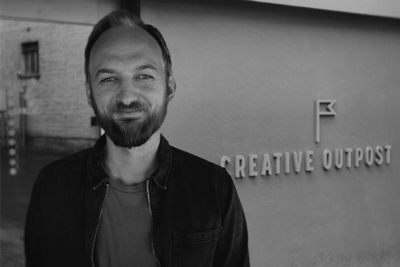Black and white portrait of a smiling man with a beard standing in front of a wall labeled Creative Outpost.