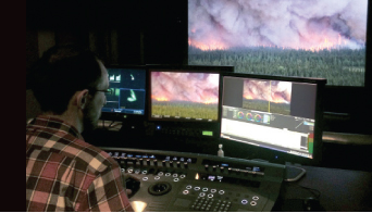 A person in a plaid shirt sits before several monitors displaying footage of a forest fire.