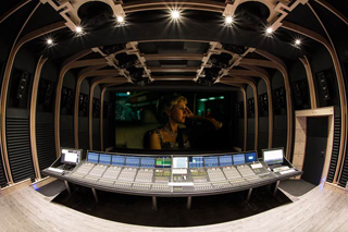 A wide view of a professional audio mixing suite with a large console and a movie playing on a screen.