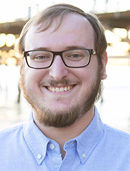 A man with brown hair, glasses, and a beard smiling in a light blue button-down shirt.