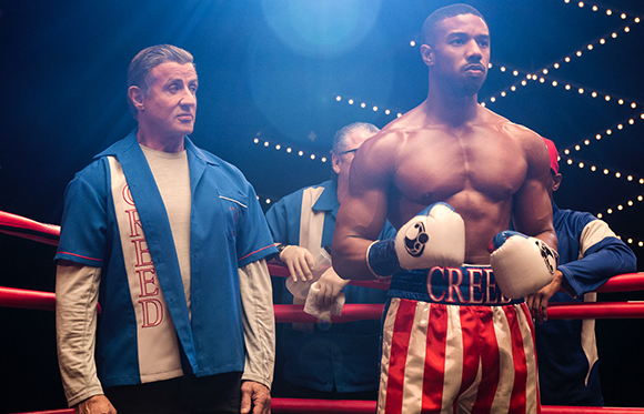 A shirtless boxer in red and white trunks stands next to a trainer in a blue shirt in a boxing ring.