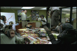 Several chimpanzees wearing clothes act out a chaotic scene in a modern office around a conference table.