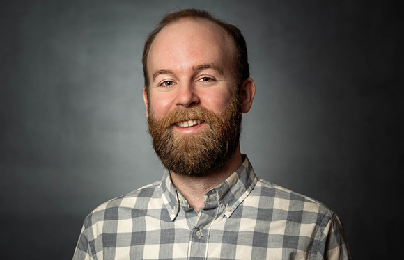A smiling man with a brown beard and receding hairline wearing a grey and white checkered button-down shirt.