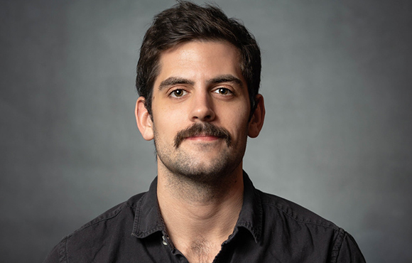 A man with dark hair and a thick mustache looking directly at the camera against a gray background.