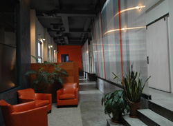 A hallway featuring orange leather armchairs, potted plants, and large glass panels along a concrete floor.