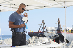 A man in a blue t-shirt and sunglasses talks on a phone while holding a white object on a beach.