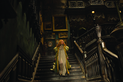 A person in a long green and cream period dress walks down a dark, ornate wooden staircase in a mansion.