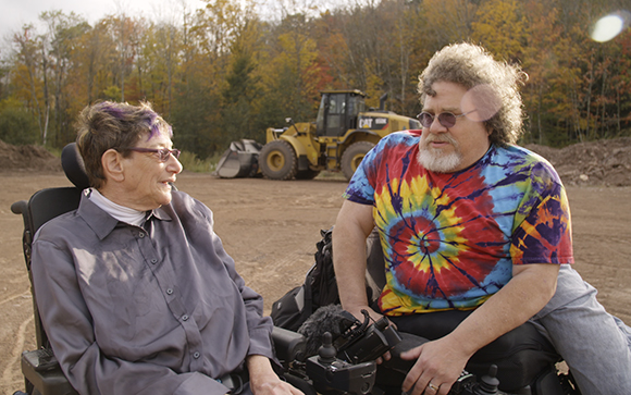 A woman and a man in wheelchairs sit outdoors and talk near a yellow tractor.