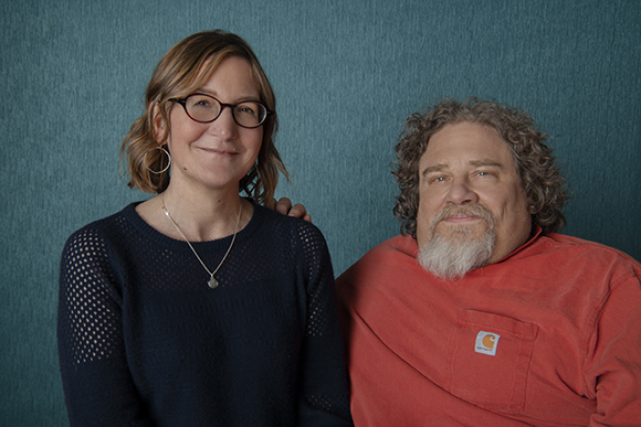 A woman with glasses and a man with a grey beard pose together against a textured teal wall.