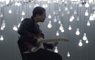 A man sits on a chair playing an electric guitar against a backdrop of many hanging glowing light bulbs.