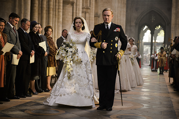 A bride in a white lace wedding dress walks down a cathedral aisle escorted by a man in a black naval uniform.
