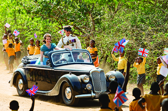 A man in a white military uniform and a woman in a blue dress drive a black convertible past cheering children.