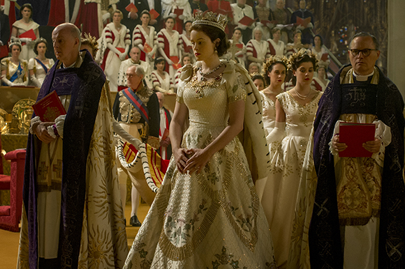 A woman in a white royal gown and crown stands in a cathedral surrounded by clergy and people in formal attire.