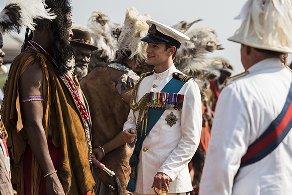 A man in a white military uniform with a blue sash and medals talks to people in traditional indigenous clothing.