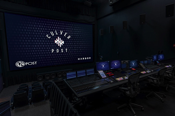 A dark post-production suite featuring a large screen displaying a logo and multiple audio mixing consoles.