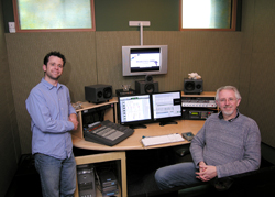 Two men posing in a professional audio recording studio with monitors and mixing equipment.