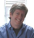 A smiling man with light brown hair wearing a blue button-down shirt.