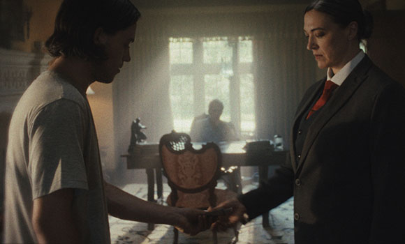 A man in a t-shirt and a woman in a dark suit stand facing each other in a sunlit room.
