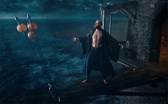 A shirtless man in dark robes stands on a wooden platform overlooking a dark, stormy ocean.