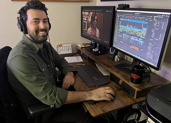 A man with headphones smiles while sitting at a desk with two computer monitors displaying video editing software.