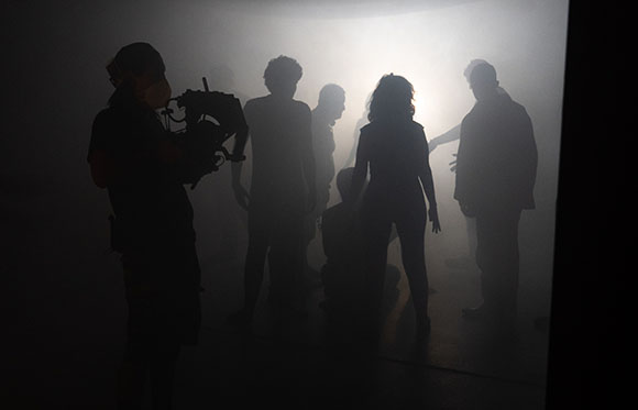 Silhouettes of several people standing in a thick, bright fog with a camera operator in the foreground.