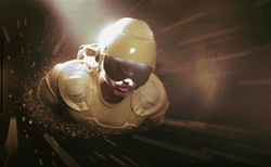 A person in tan futuristic armor and a helmet moving through a dark, streaked tunnel.
