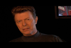 A man with styled brown hair wearing a dark turtleneck against a black background.