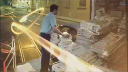 A man stands beside large piles of newspapers on a street at night with light trails passing by.