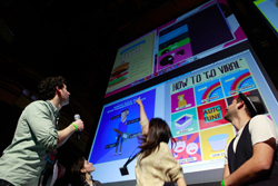 A group of people looking up at a large presentation screen displaying colorful graphics and text.
