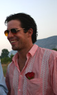 A smiling man with dark hair wearing sunglasses and a pink embroidered shirt with a red rose in his pocket.