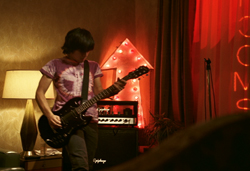 A person playing a black electric guitar in a room with warm and red lighting.