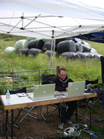 A person sits at a wooden table using two laptops under a white canopy tent in a grassy field.