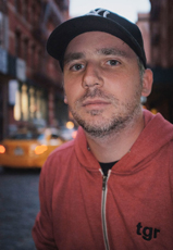 A man wearing a black baseball cap and a red zip-up hoodie stands on a city street.